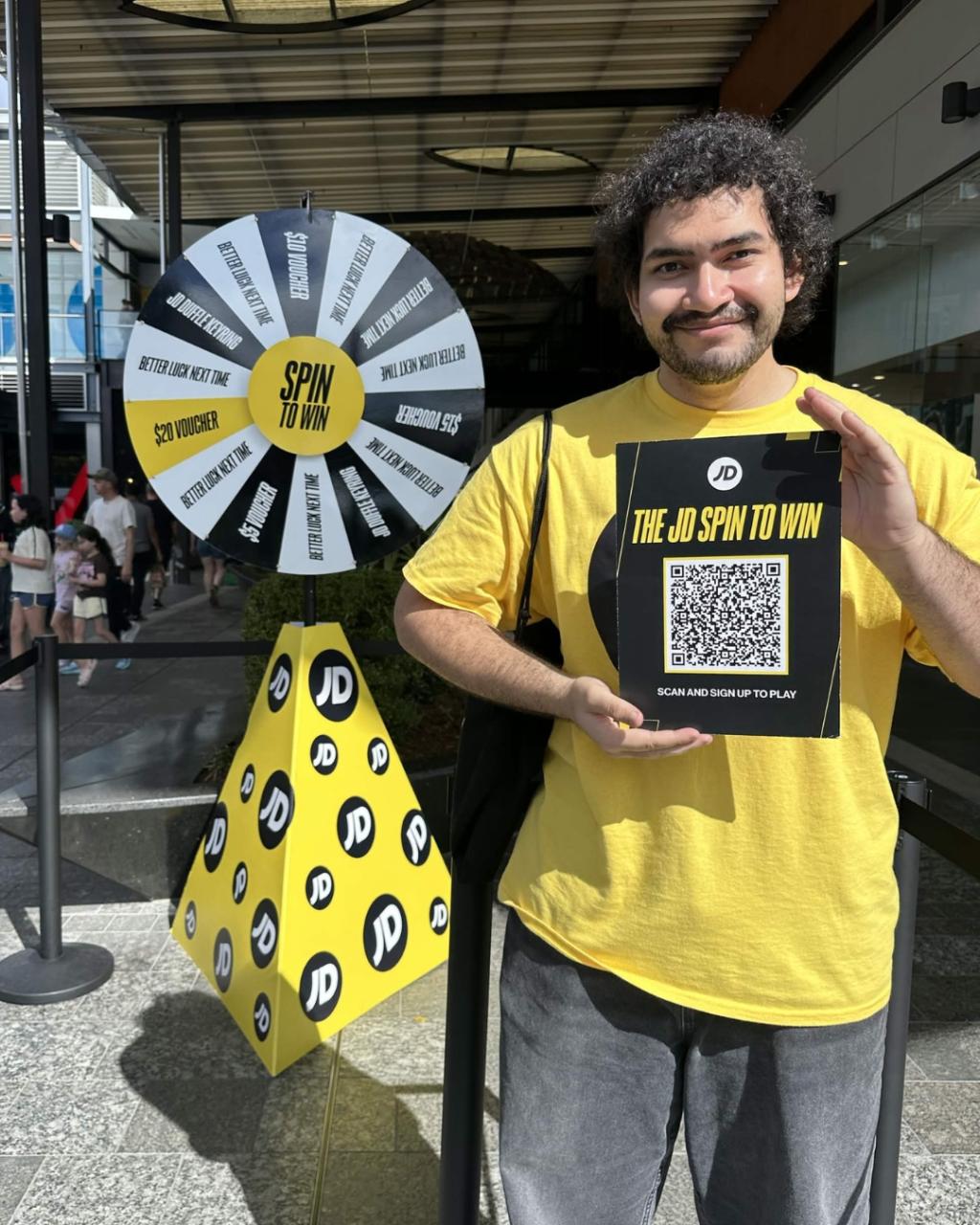 Male brand ambassador in JD Sports branded uniform giving a thumbs up beside the Coin Drop activation wheel